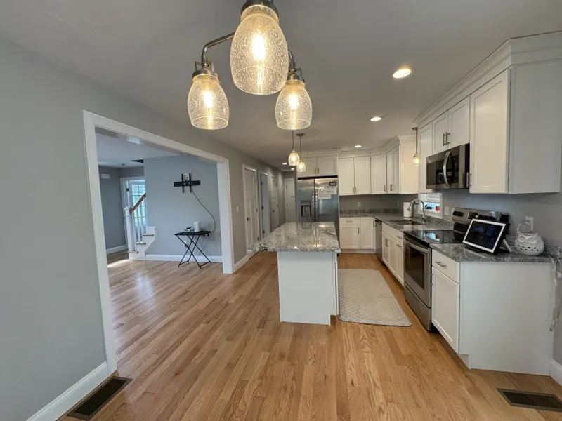 Modern kitchen with white cabinets, granite island, and hanging pendant lights, wood floors.