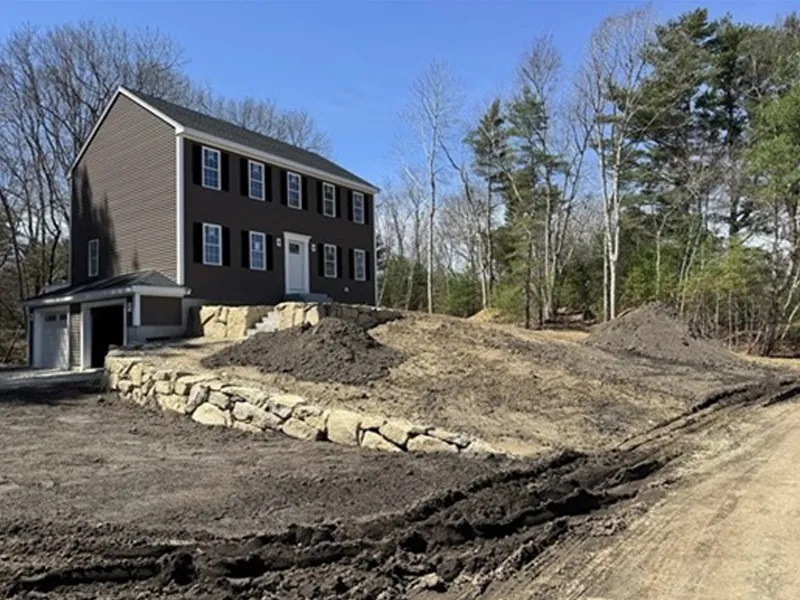 Brown two-story house with garage and dirt landscaping on a sloped, partially wooded lot.