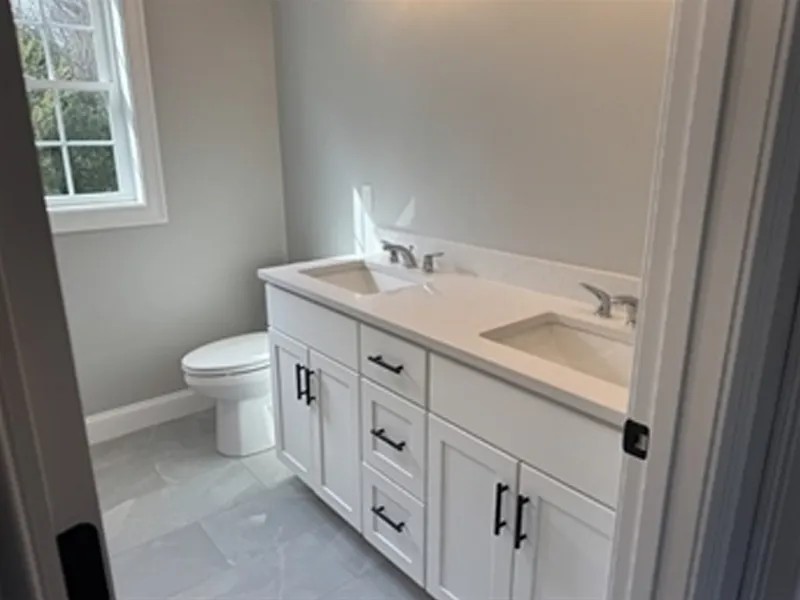 Modern bathroom with double sinks, white cabinets, and a toilet near a window.