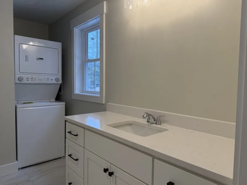 Laundry area with stacked washer and dryer next to a sink and countertop in a bright room.
