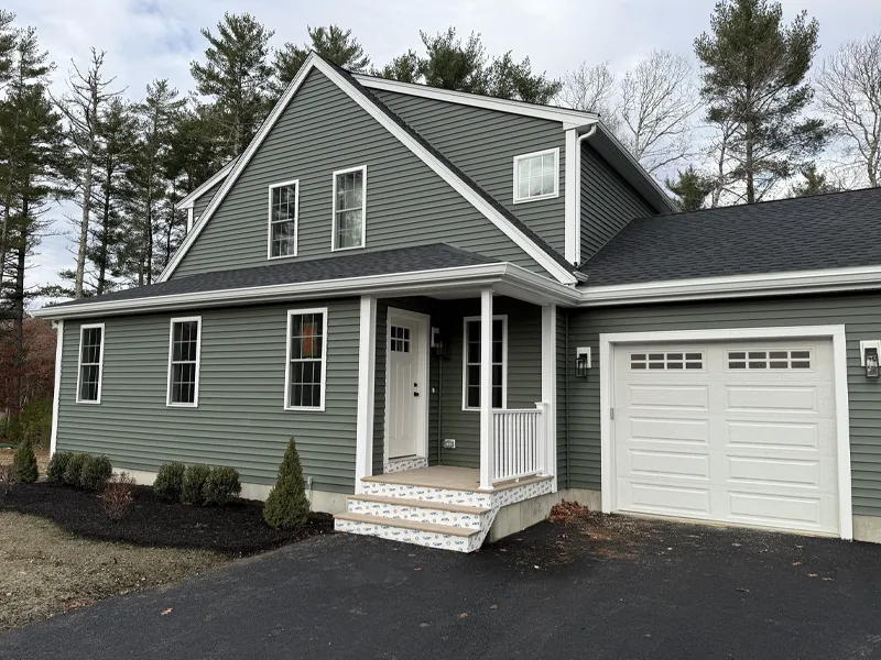 Green two-story house with a front porch, attached garage, and surrounding trees.