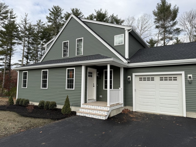Green two-story house with white trim, front porch, and attached garage in a wooded area.