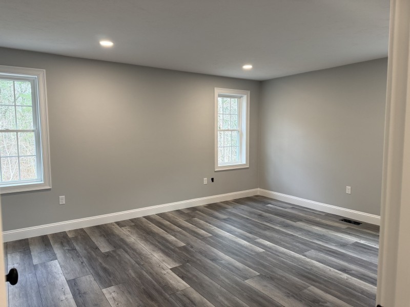 Empty room with gray walls, wood flooring, two windows, and recessed ceiling lights.