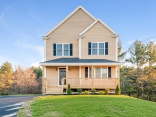 Two-story beige house with front porch, black shutters, and a green lawn on a sunny day.