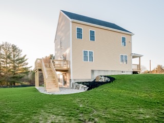 Light tan two-story house with deck and stairs, surrounded by green grass and trees.