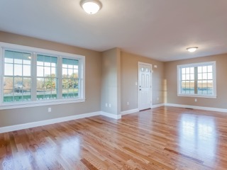 Bright, empty room with hardwood floors, beige walls, and large windows letting in sunlight.