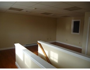 Empty room with beige walls, wood floor, window, and partial stair railing in sunlight.