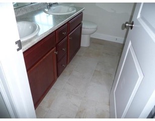 Bathroom with double sinks, wooden cabinets, tiled floor, and a white toilet.