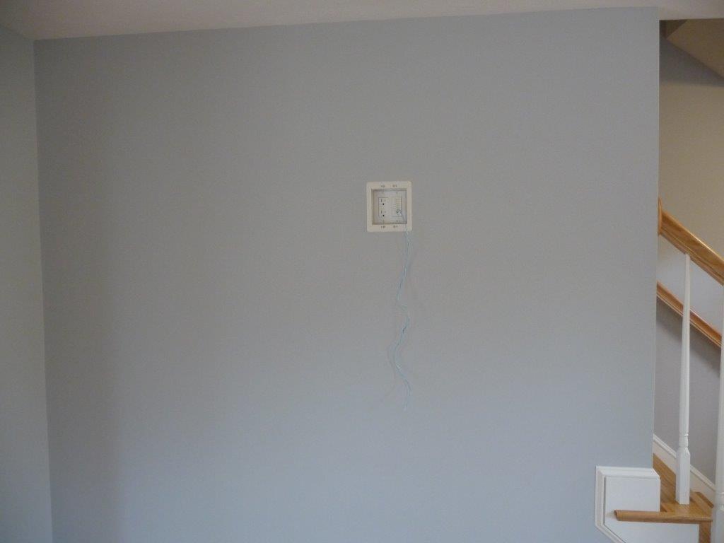 Light gray wall with a small wall panel and hanging blue wires, next to a stair railing.