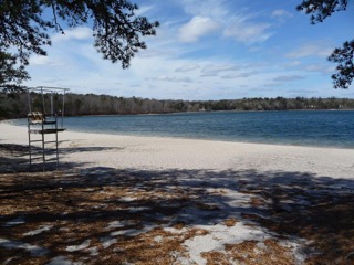 Sandy beach with clear blue water, empty lifeguard stand, and trees under a partly cloudy sky.
