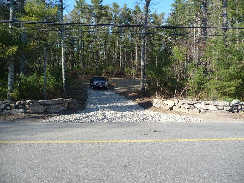 Black truck parked on gravel driveway leading from a forest to a paved road.