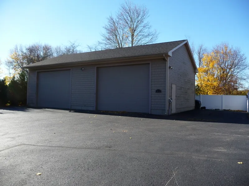 Large gray garage with three closed doors on an asphalt lot, trees in the background.
