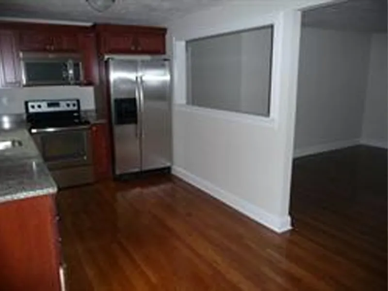 Kitchen with stainless steel appliances and wood floors, next to an empty, open room.