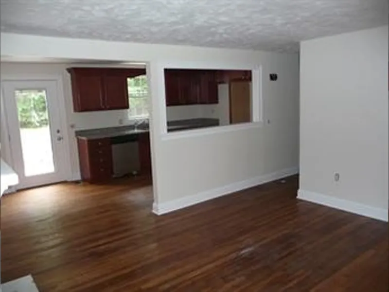 Empty living room with hardwood floors and a view into a kitchen with dark cabinets.