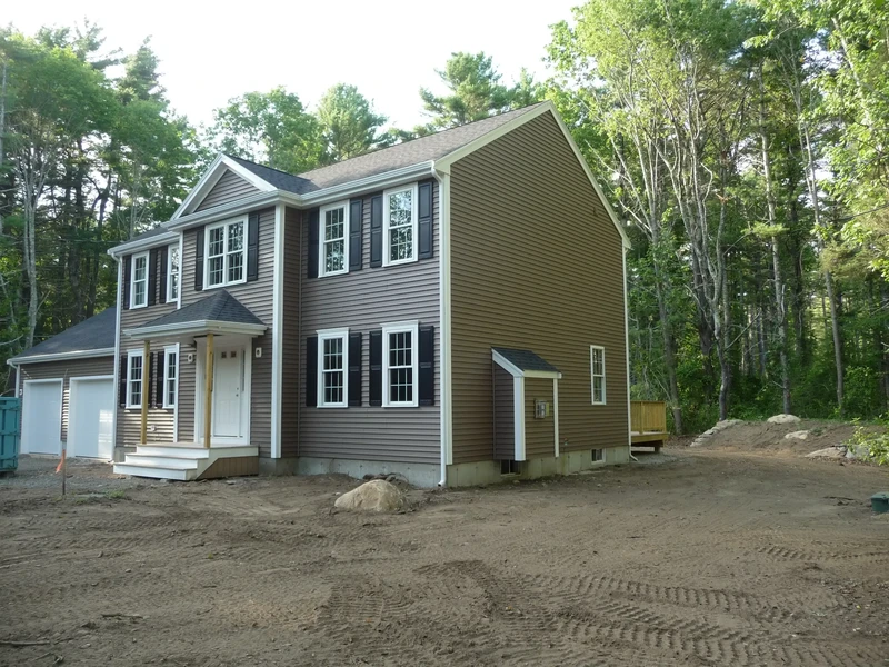 Two-story brown house with white trim and black shutters, surrounded by trees and dirt yard.
