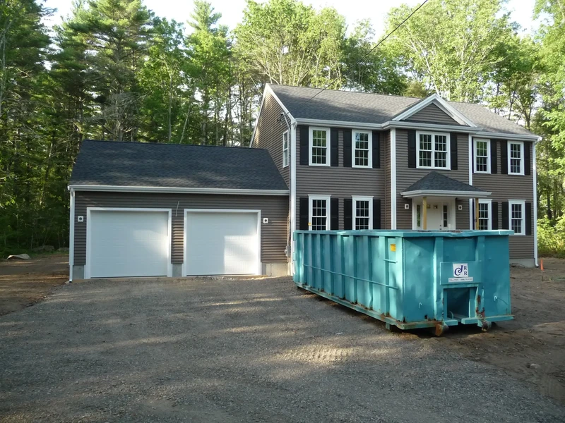 Two-story house with double garage and large blue dumpster in the gravel driveway.