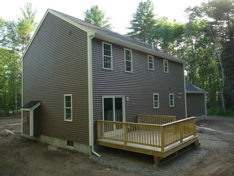 Two-story brown house with a wooden deck in the backyard, surrounded by trees.