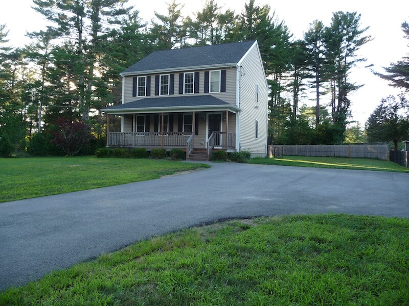 A two-story house with a front porch, driveway, and trees in the background.