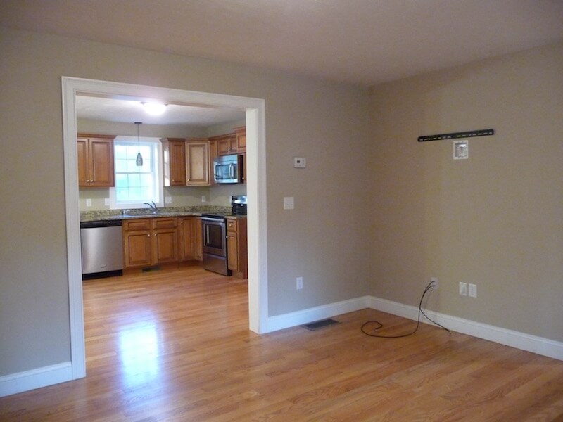 Open kitchen with wooden cabinets, seen from an empty room with hardwood floors.