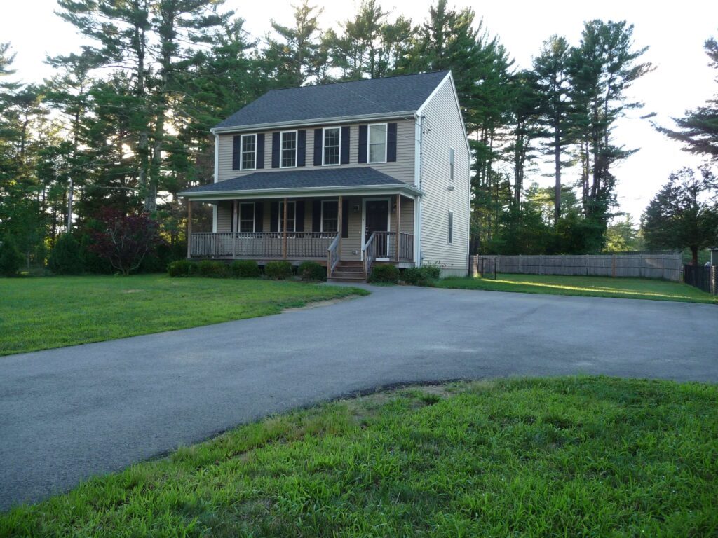 Two-story beige house with a front porch, large yard, and trees in the background.