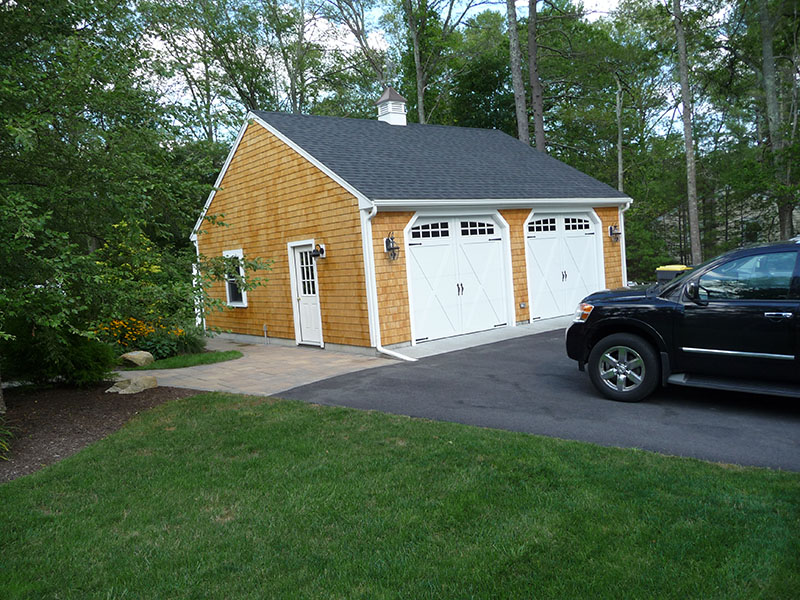 A tan detached garage with two white doors next to a black SUV and green lawn.