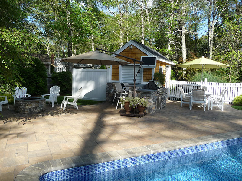 A backyard patio with a pool, fire pit, patio chairs, umbrellas, and a small pool house.