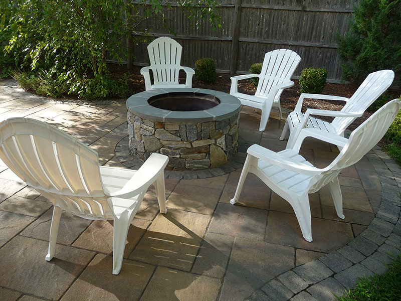 Five white plastic chairs surround a stone fire pit on a paved patio in a backyard.