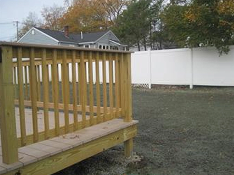 Small wooden deck in a backyard with grass, white fence, and a house in the background.