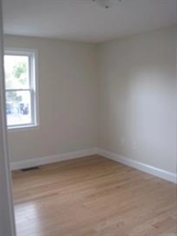 Empty room with light-colored walls, wood floor, and a single window on the left wall.