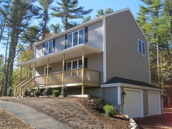 Two-story beige house with a porch, double garage, and surrounded by tall trees.