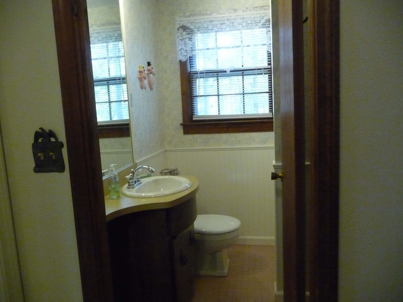 A small bathroom with a sink, toilet, mirror, and a window with blinds and a lace curtain.