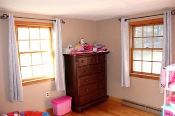 A dresser with toys on top is between two windows in a sunlit, tidy room.