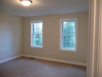 Empty room with beige walls, two windows, and carpeted floor. Ceiling light is on.
