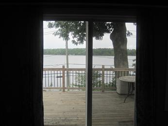 View through glass doors of a wooden deck overlooking a lake with trees in the background.