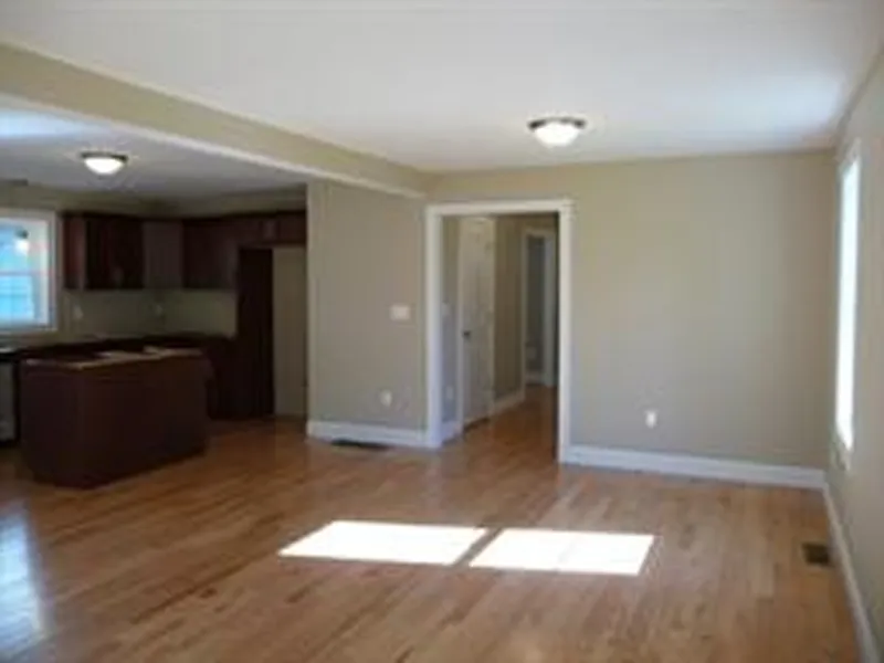 Empty living room with hardwood floors, beige walls, and view of a kitchen in the background.