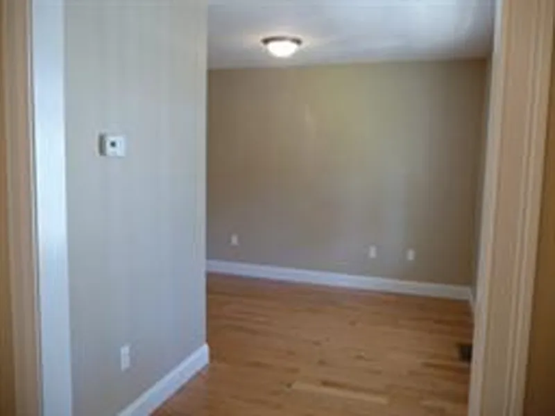 Empty beige room with light wood floor, white trim, and a ceiling light fixture.