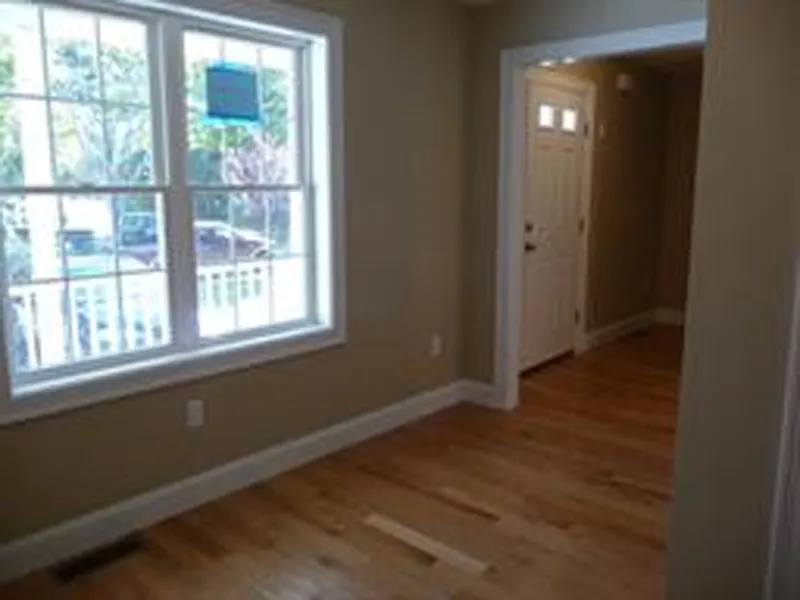 A small empty room with hardwood floors, beige walls, and a window near a white front door.