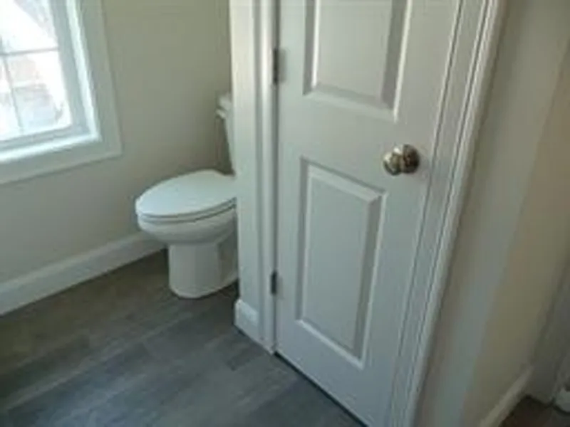 A white door partially blocks access to a toilet in a small bathroom with a window.