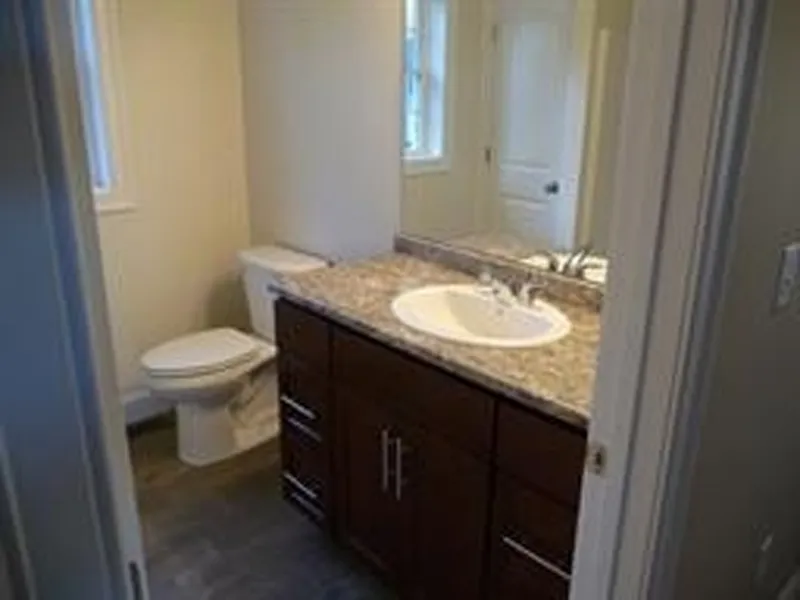 Small bathroom with a toilet, sink, mirror, and dark wood vanity under bright lighting.