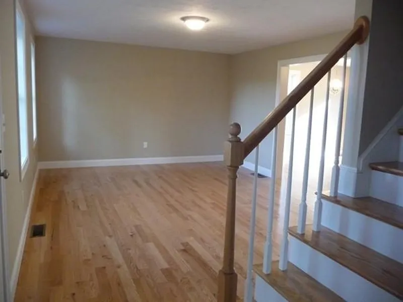 Empty living room with hardwood floors, beige walls, and a staircase with wooden railing.