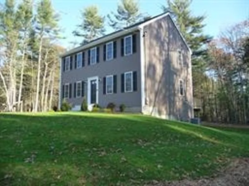 Two-story gray house with black shutters on a grassy lawn, surrounded by tall trees.