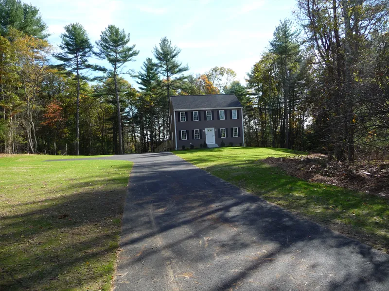 A paved driveway leads to a two-story house surrounded by trees and a grassy yard.