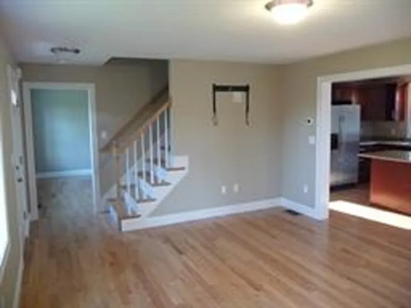 Empty room with wooden floors, a staircase, and view of a kitchen with stainless appliances.