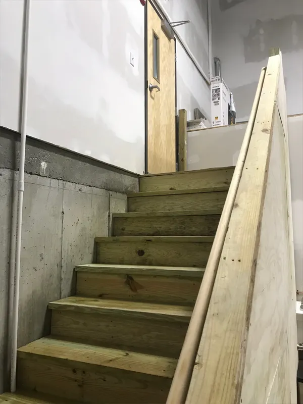 Wooden stairs leading up to a door mounted unusually high on a wall in an unfinished room.