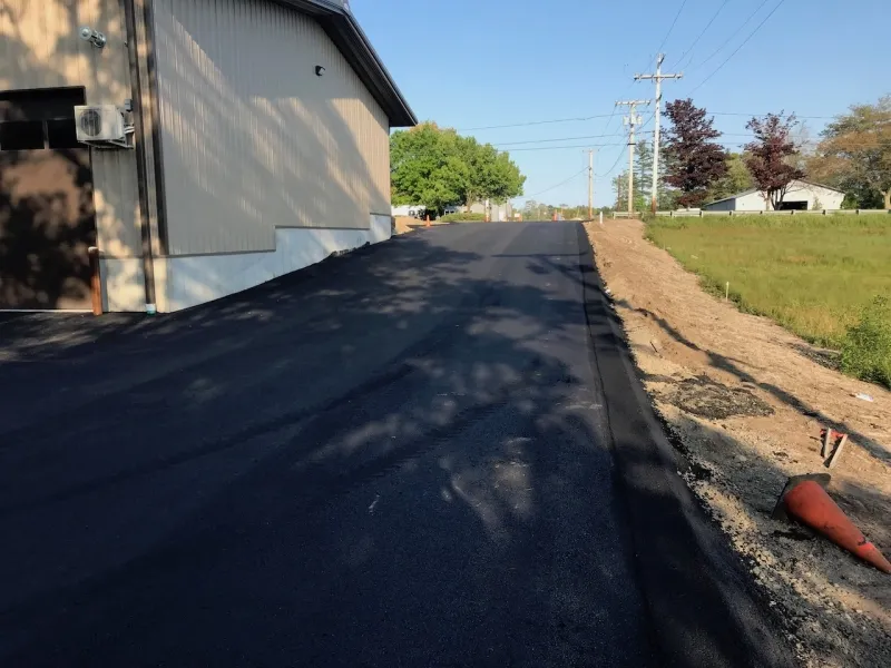 Freshly paved asphalt driveway beside a building, with grass and trees on the right side.