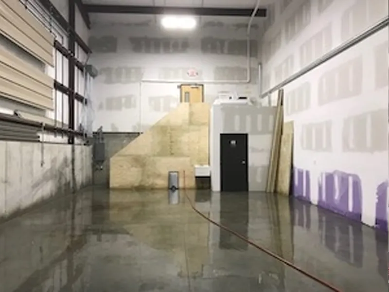Warehouse with wet, shiny concrete floor, unfinished drywall, and wooden stairs in back.