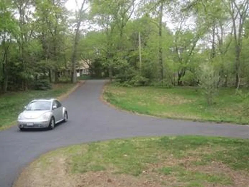 A silver car drives on a paved road curving through a green, wooded area near a house.