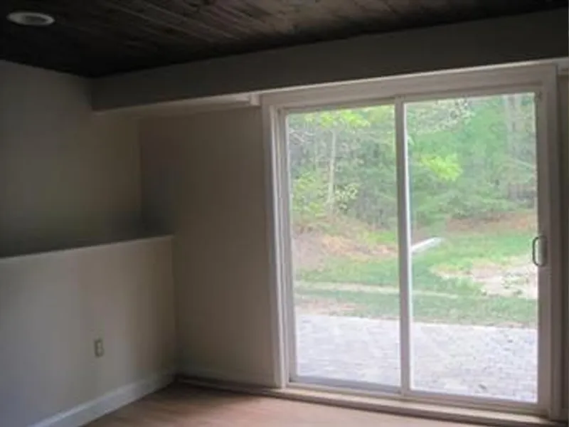 Room with wood floor, white walls, and large sliding glass door overlooking greenery outside.