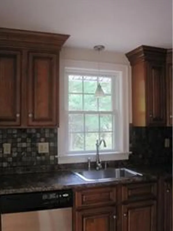 Kitchen sink under a window with wooden cabinets, dark countertop, and tile backsplash.