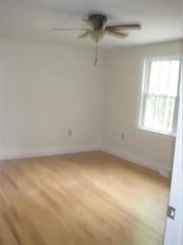 Empty room with hardwood floors, white walls, a ceiling fan, and a window with sunlight.